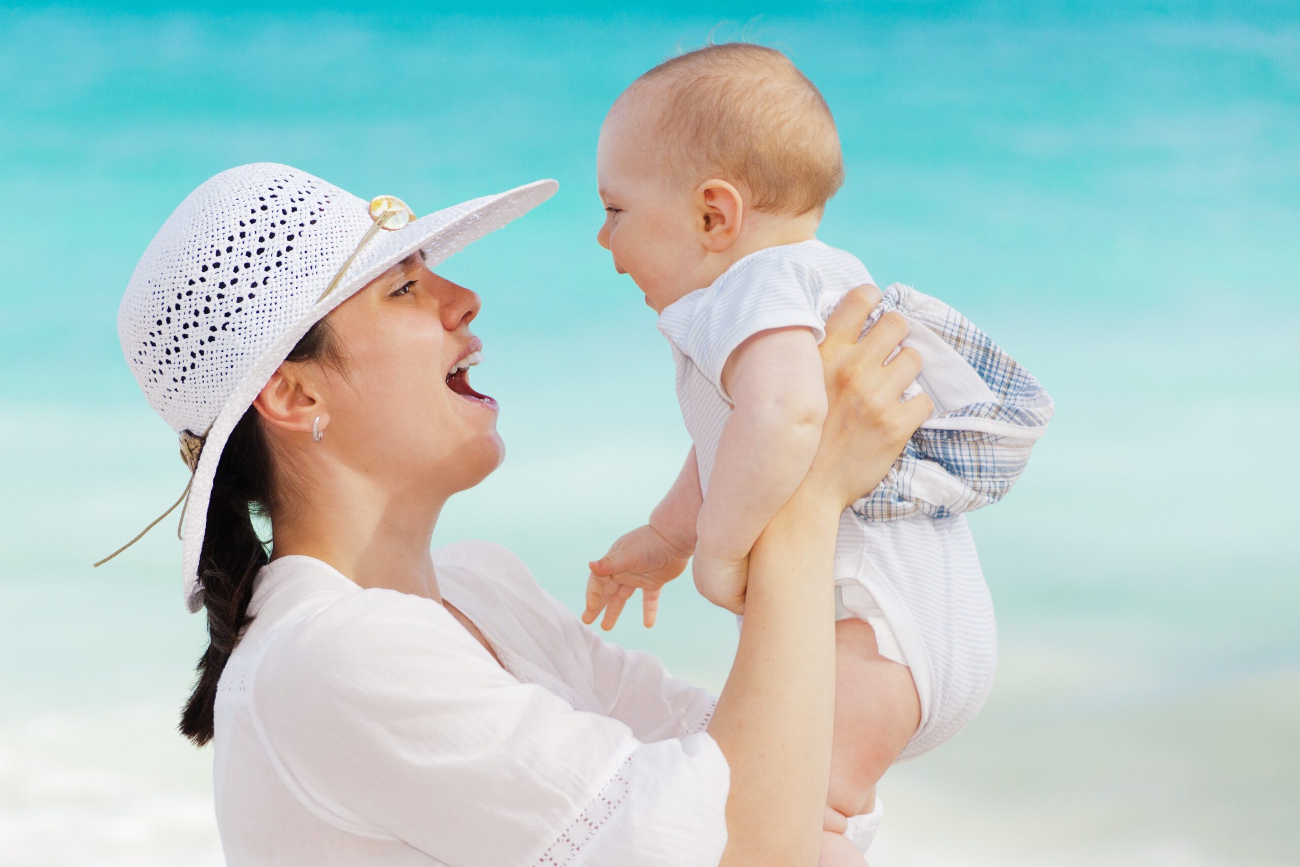 Woman in a white sun hat lifts a baby against a turquoise beach backdrop.